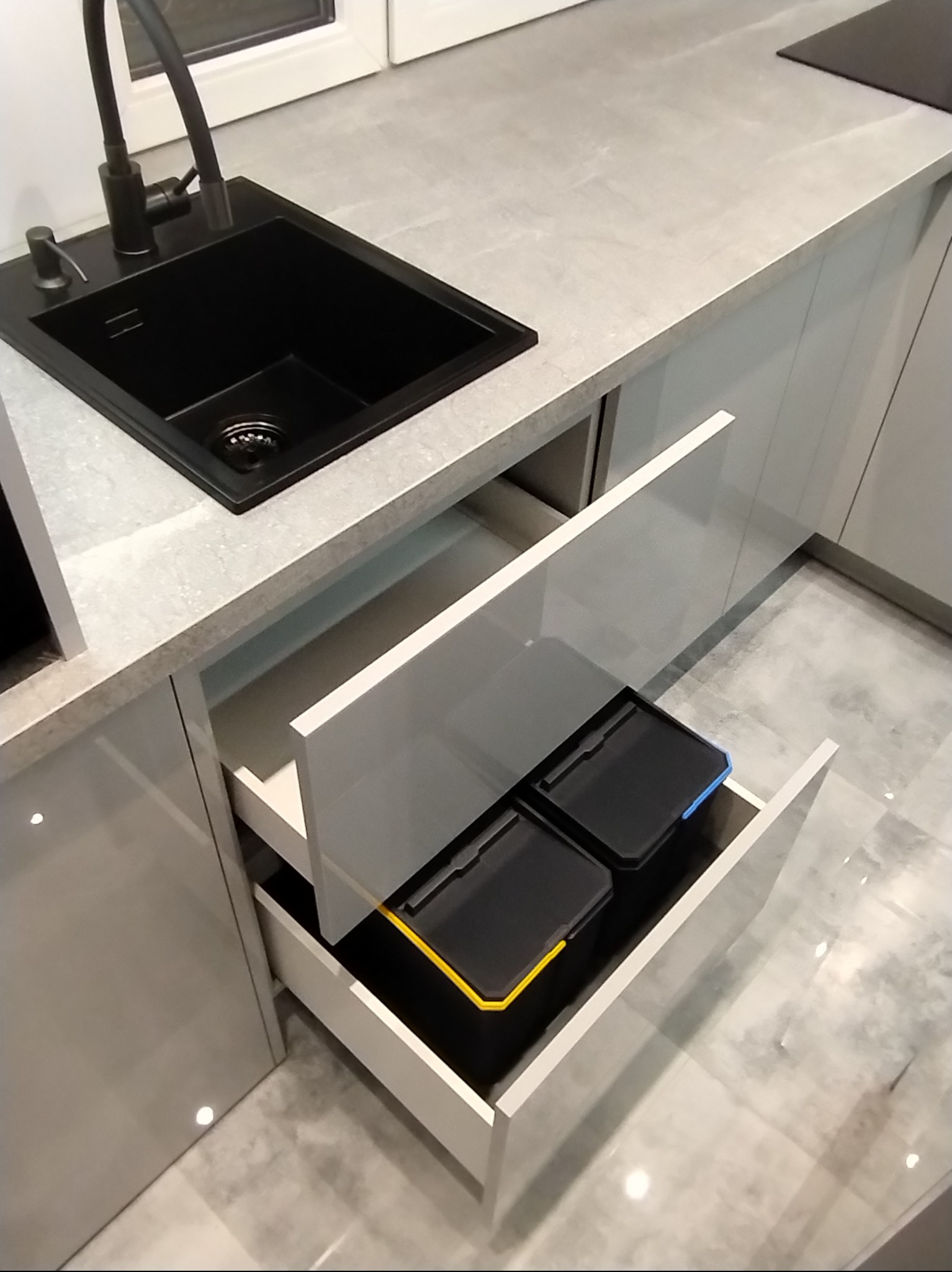 Nowoczesna kuchnia z szarymi frontami, czarnym zlewem i baterią. Otwarta szuflada z pojemnikami na segregację śmieci z żółtymi i niebieskimi akcentami.