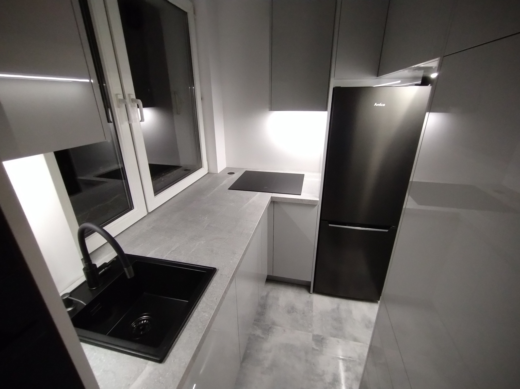 Nowoczesna kuchnia w szarościach: czarny zlew z baterią, szary blat, białe szafki i czarna lodówka Amica. Minimalistyczny design, oświetlenie LED pod szafkami.