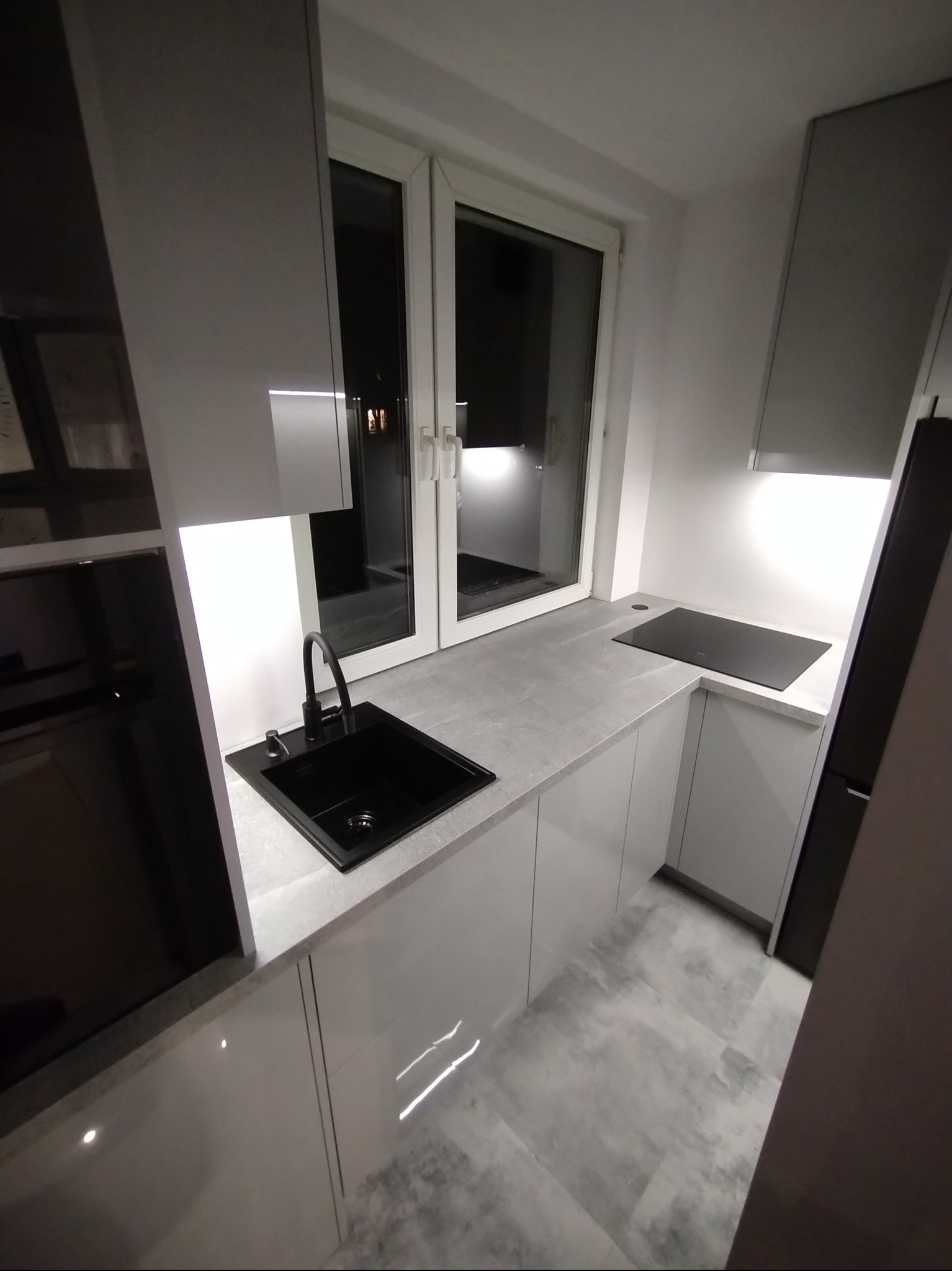 Nowoczesna kuchnia z czarnym zlewem i płytą indukcyjną, szare blaty, białe szafki z połyskiem, oświetlenie LED pod szafkami, okno w tle. Minimalistyczny design.