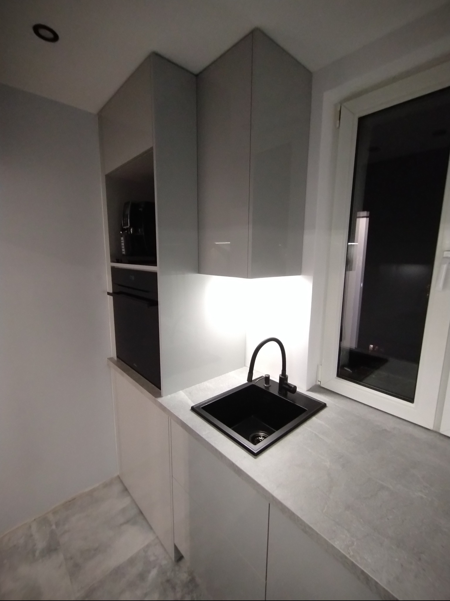 Nowoczesna kuchnia z szarymi szafkami, czarnym zlewem i armaturą. Podświetlenie LED pod szafkami górnymi. Minimalistyczny design, widok na okno.