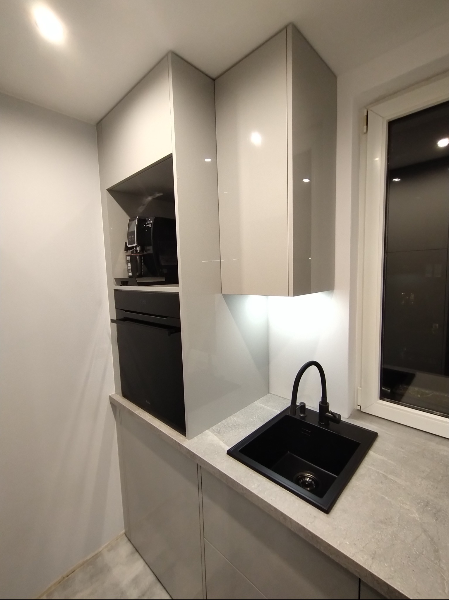 Nowoczesna kuchnia: białe szafki z połyskiem, czarny zlew i bateria, wbudowany piekarnik i ekspres do kawy. Minimalistyczny design, oświetlenie LED pod szafkami.