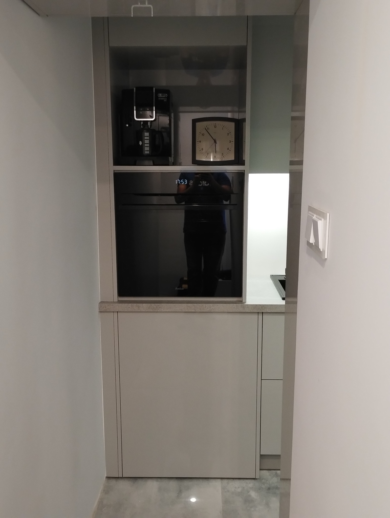 Szare szafki kuchenne z wbudowanym piekarnikiem Amica, ekspresem do kawy i zegarem. Minimalistyczny design, nowoczesne oświetlenie, widok z perspektywy korytarza.