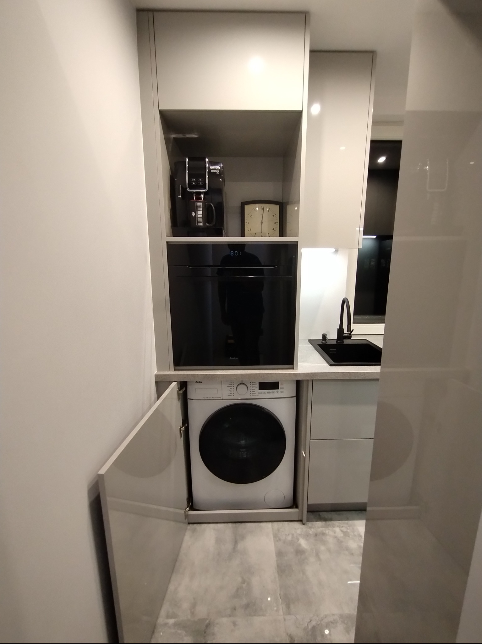 Nowoczesna kuchnia z zabudową: pralka, piekarnik, ekspres do kawy. Jasne fronty, czarny zlew i bateria. Minimalistyczny design, szara podłoga.