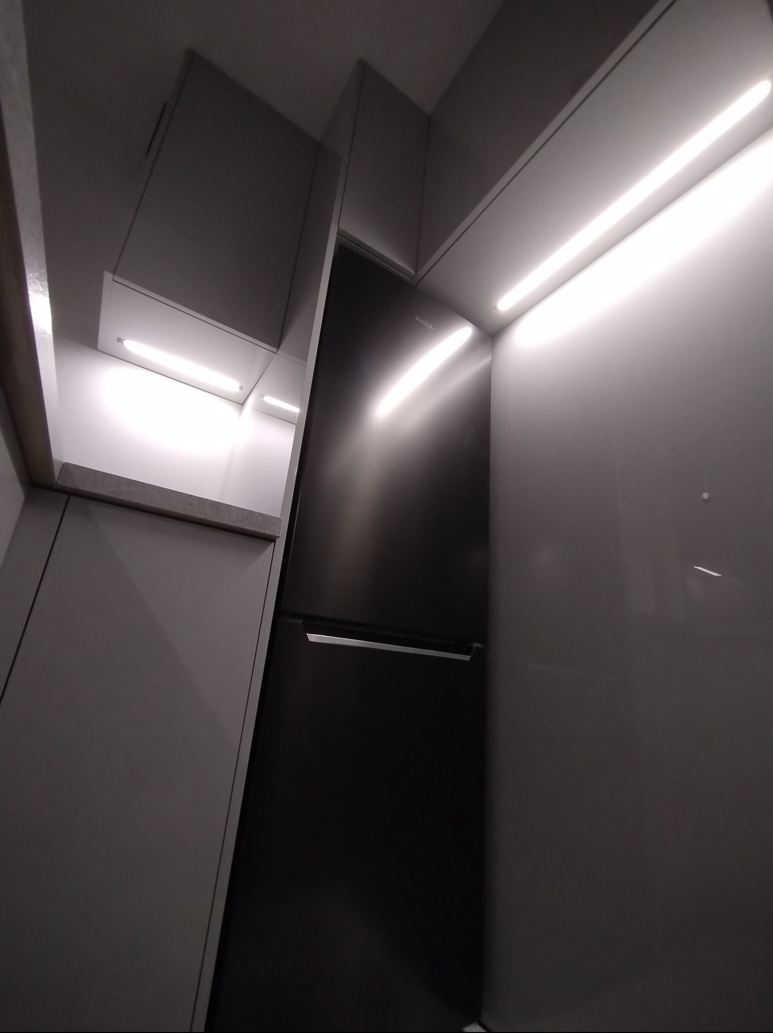 Nowoczesna, szara zabudowa kuchenna z oświetleniem LED i czarną lodówką, sfotografowana pod nietypowym kątem, ukazująca minimalistyczny design.