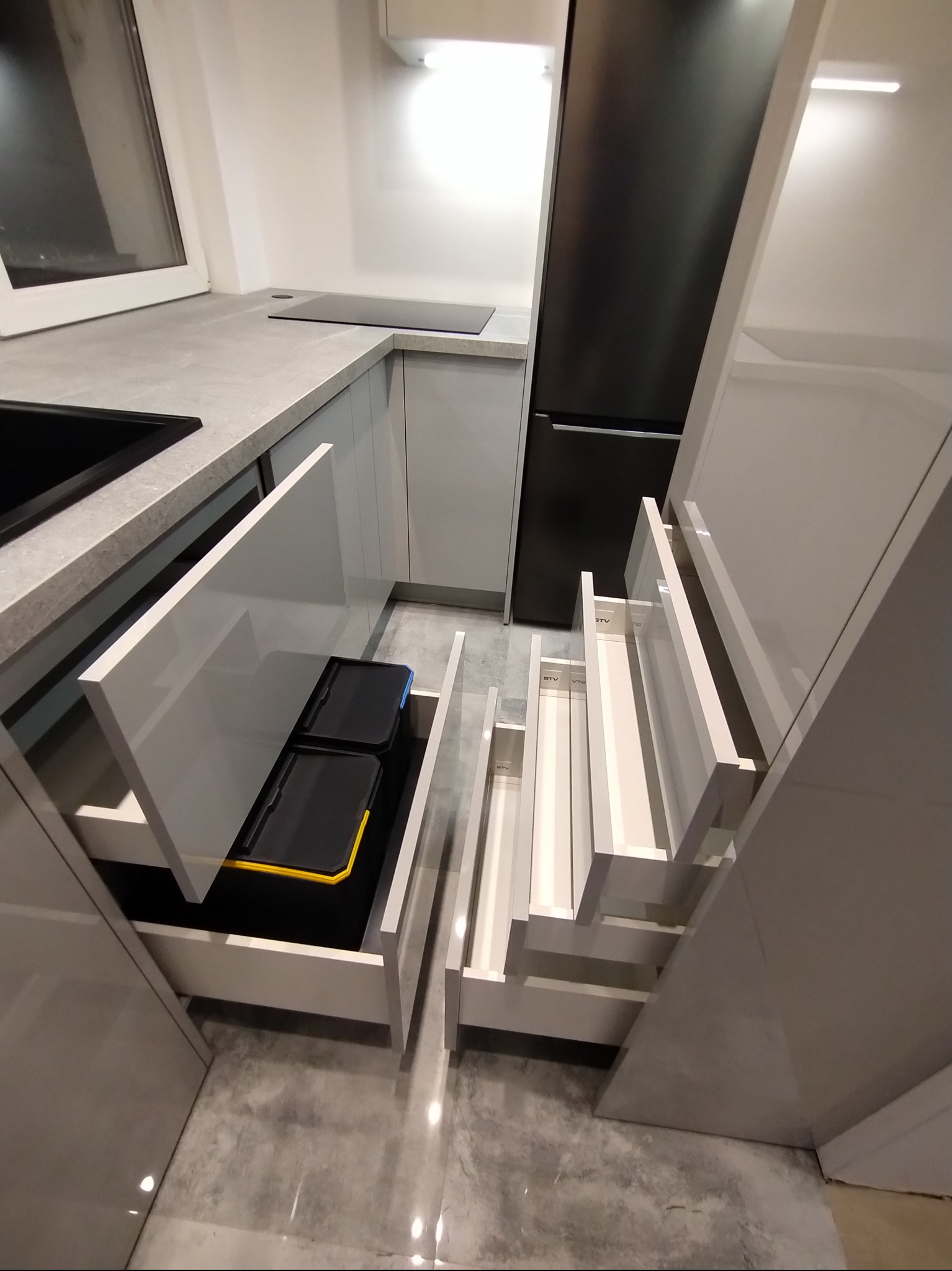 Nowoczesna kuchnia z szarymi frontami, otwartymi szufladami i koszem na śmieci. Widoczny blat i płyta indukcyjna. Minimalistyczny design, oświetlenie LED.