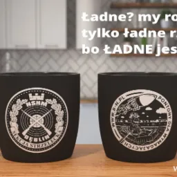 Dwa czarne kubki z białymi nadrukami, jeden z logo sekcji strzeleckiej Dęblin, drugi z motywem pontonów dla wymagających, ustawione na drewnianym blacie w tle kuchni.