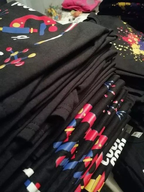 Stos czarnych koszulek z kolorowymi nadrukami, z bliska, na stole. Widoczne wzory: abstrakcyjne kształty, molekuły, plamy farby.