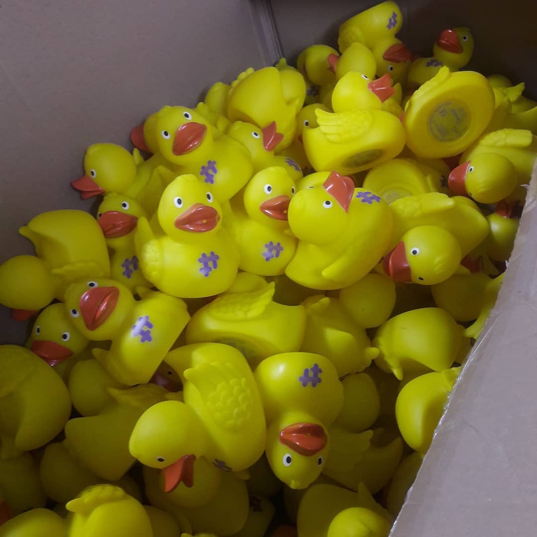 Żółte gumowe kaczuszki z fioletowymi nadrukami w kształcie krzyżyków, stłoczone w kartonowym pudełku. Widok z góry, oświetlenie naturalne.