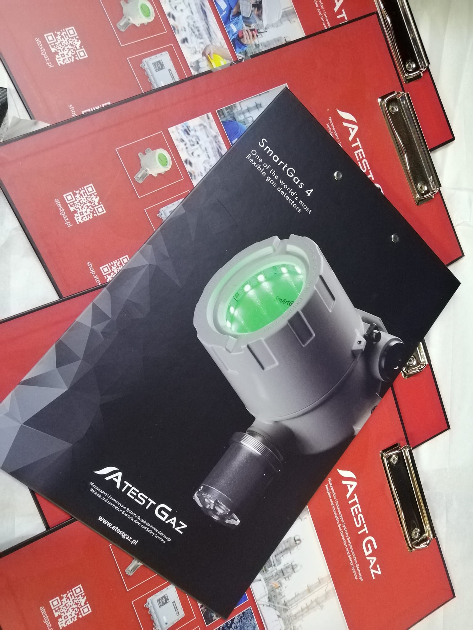 Katalogi firmowe ATEST GAZ na podkładkach z klipsem. Katalog z czarnym tłem prezentuje detektor gazu SmartGas 4. Czerwone katalogi z QR kodem i adresem www.