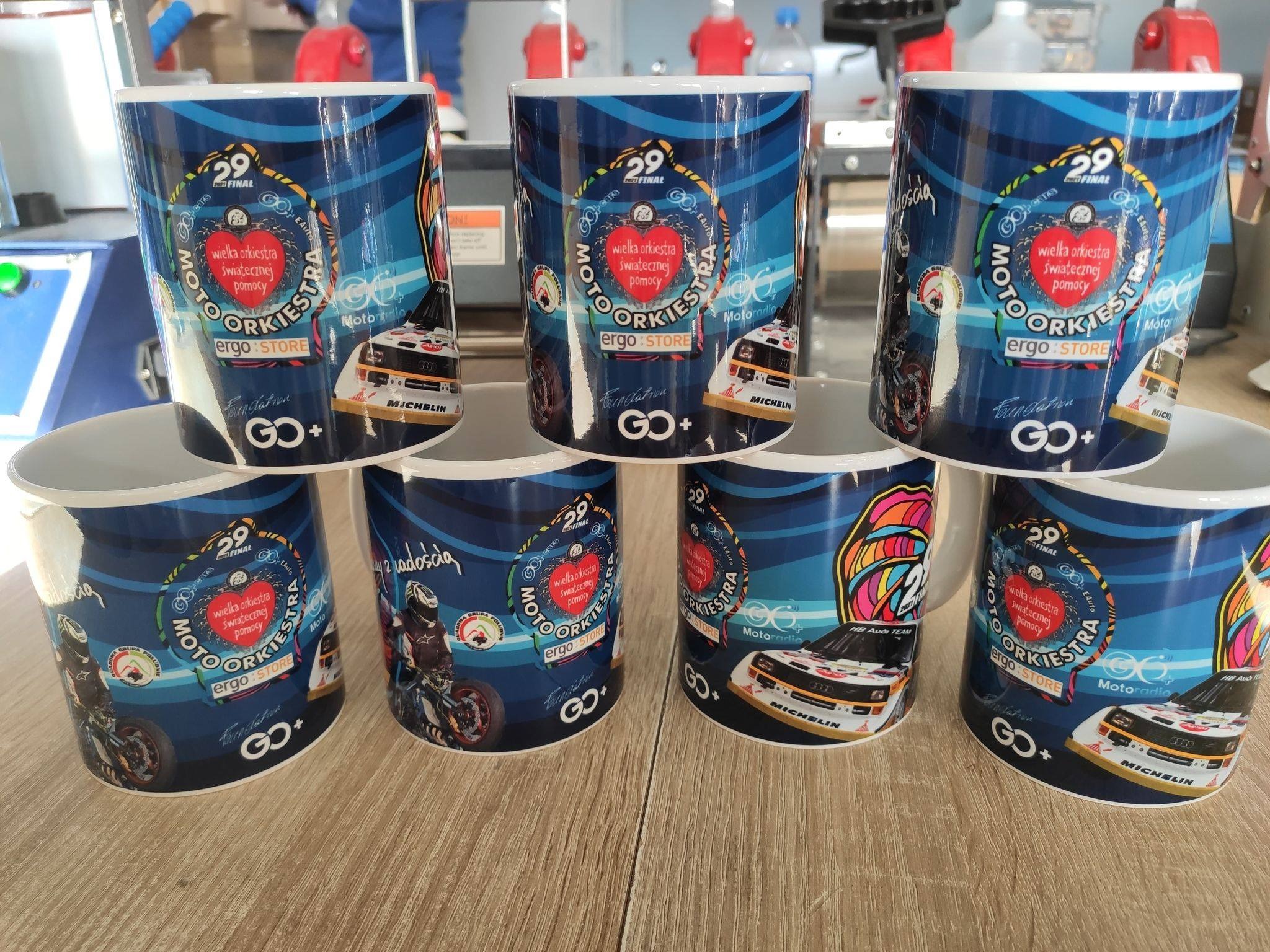 Siedem białych kubków z niebieskim nadrukiem MotoOrkiestry, ustawionych na drewnianym blacie. Widoczne logo, motywy motoryzacyjne i elementy graficzne związane z akcją charytatywną.