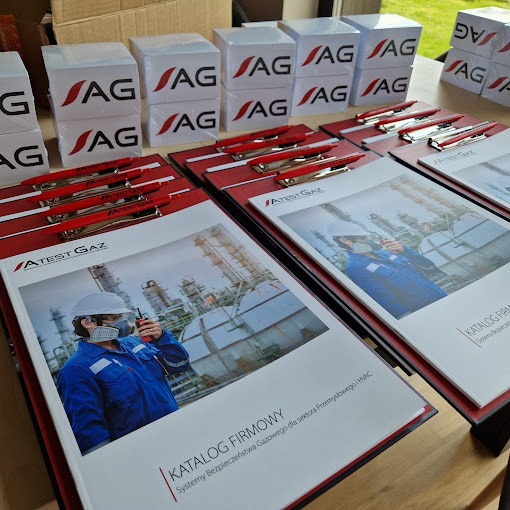 Stosy notesów i teczek z logo firmy ATEST GAZ, długopisy z nadrukiem. Materiały reklamowe z grafiką pracownika w kombinezonie na tle instalacji przemysłowej.