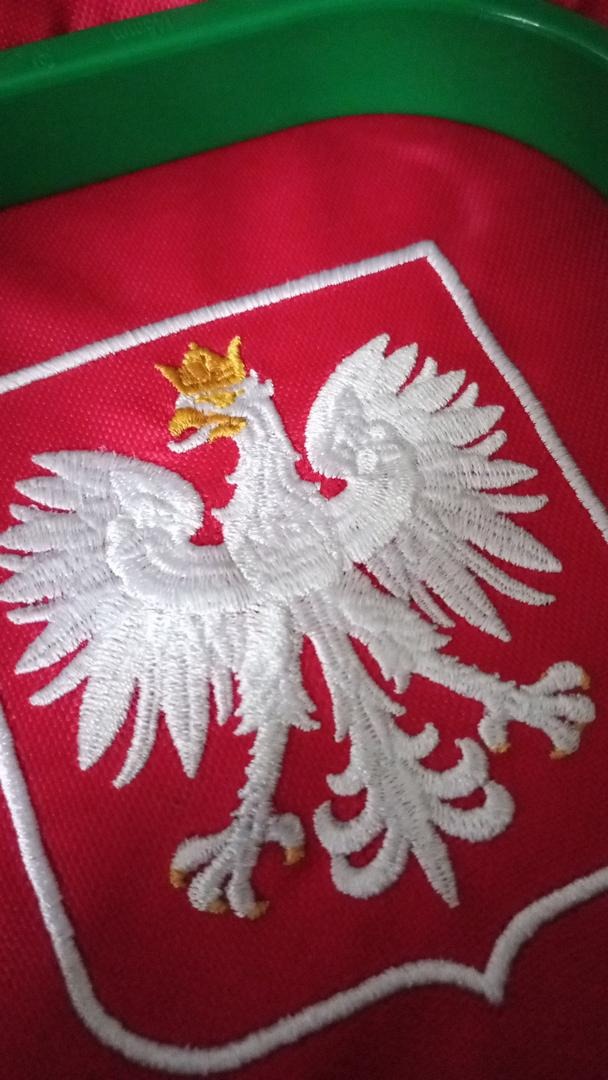 Czerwona tkanina z wyhaftowanym białym orłem w koronie, obramowanym białą nicią. Detal haftu z bliska, widoczna faktura materiału i precyzja wykonania.