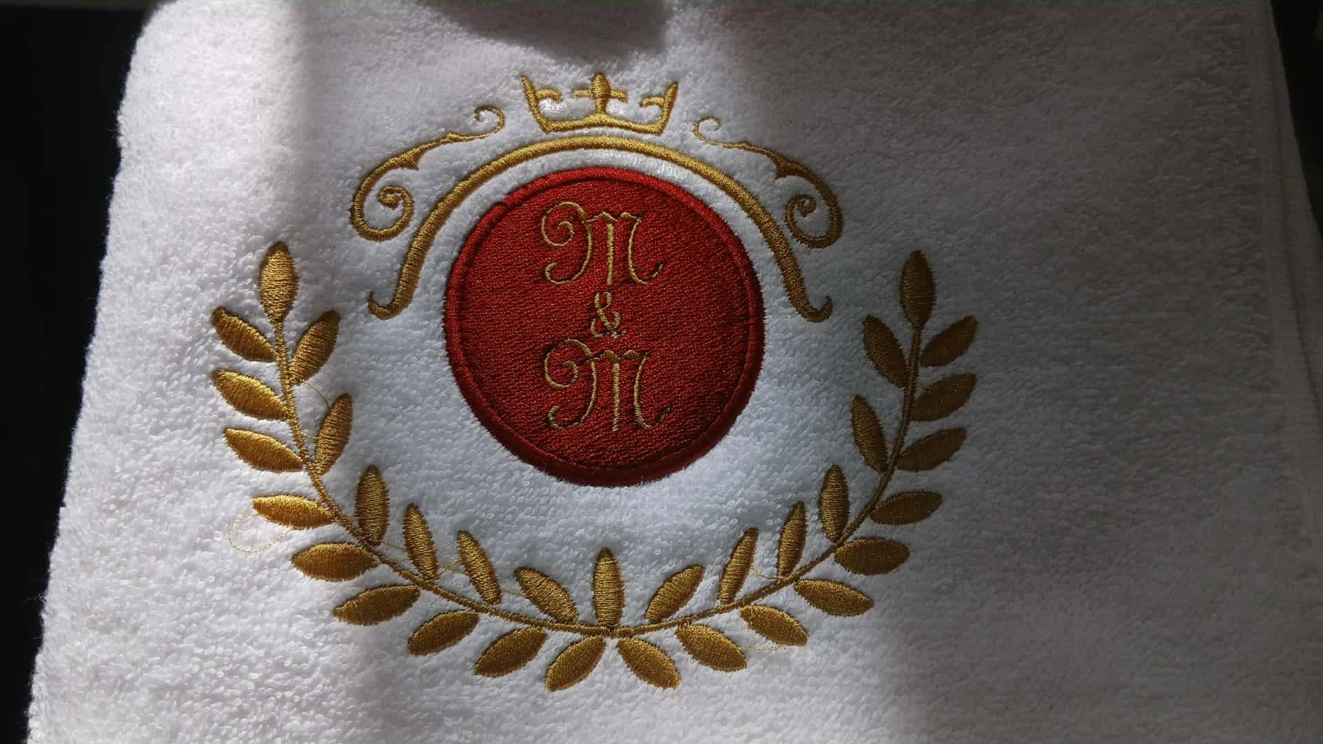 Biały ręcznik frotte z wyhaftowanym złotą nicią monogramem 'M&M' w czerwonym okręgu, otoczonym wieńcem laurowym i koroną. Elegancki detal.