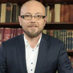 Portret mężczyzny w okularach i marynarce na tle biblioteczki z książkami.