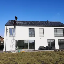 Dwóch monterów w kaskach na dachu nowoczesnego, białego domu jednorodzinnego z panelami fotowoltaicznymi, jeden wchodzi po aluminiowej drabinie, błękitne niebo w tle.