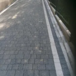 Ułożona szara kostka brukowa z białym obramowaniem, widok z perspektywy wzdłuż chodnika, obok czarne metalowe ogrodzenie.