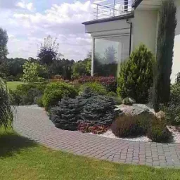 Ukształtowany ogród przydomowy z krętą ścieżką z kostki brukowej, różnorodną roślinnością ozdobną i elementami skalnymi przy jasnym budynku mieszkalnym.