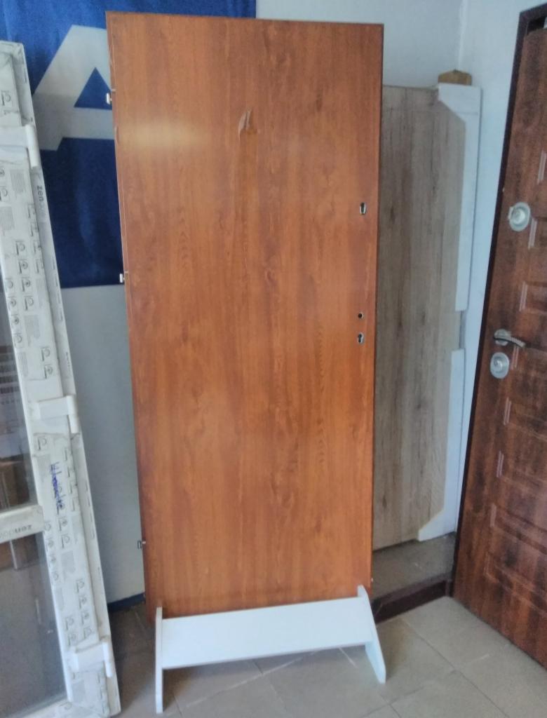 Brązowe, drewnopodobne drzwi na białym stojaku, widoczne ślady po naklejce, w tle inne drzwi i okno.