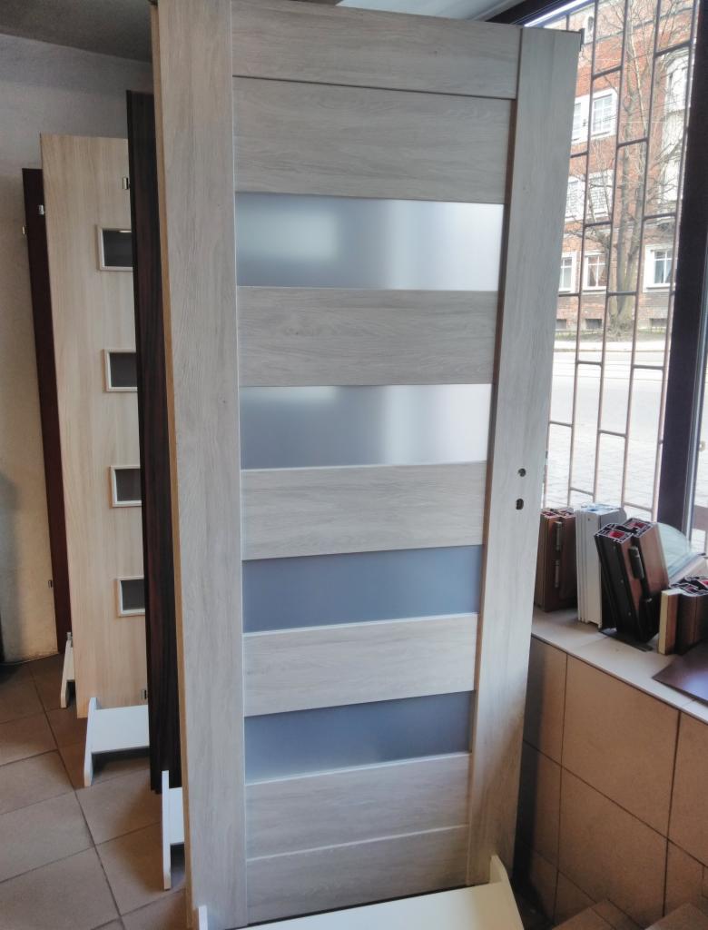 Nowoczesne, wewnętrzne drzwi z jasnego drewna z trzema poziomymi wstawkami z matowego szkła, ustawione w ekspozycji obok innych modeli drzwi.