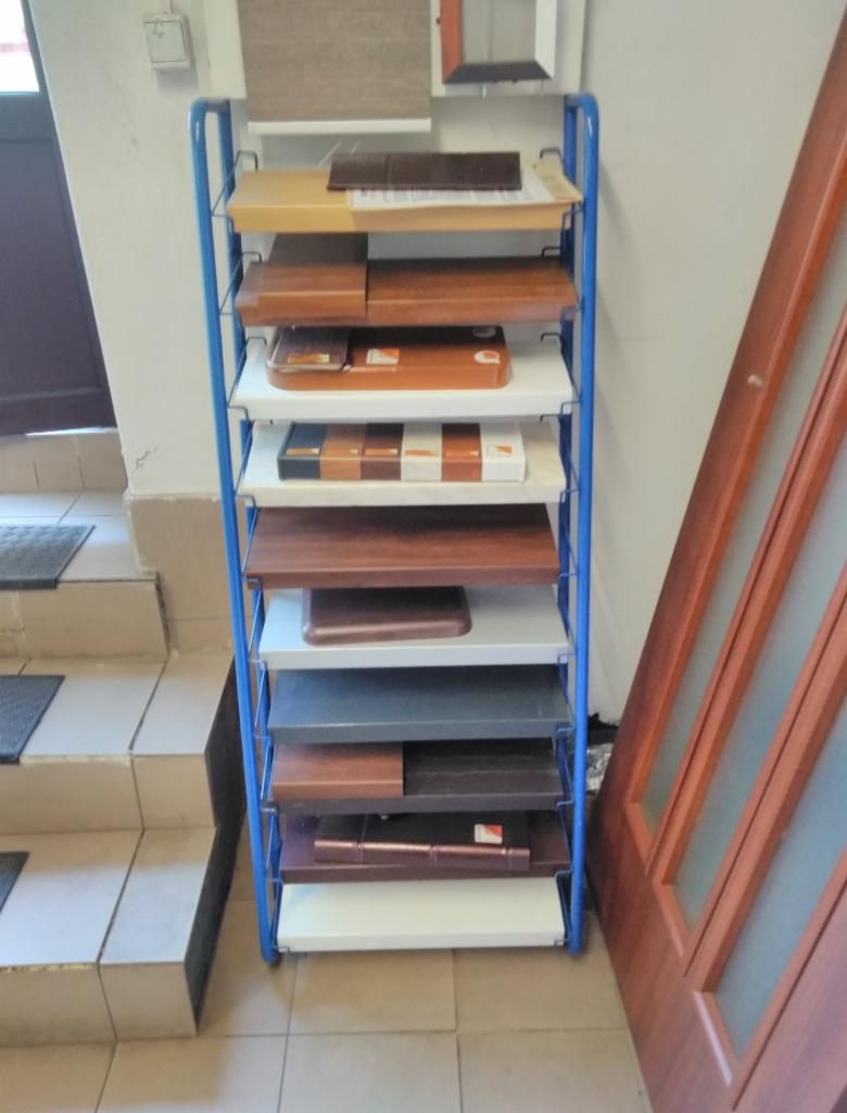 Ekspozytor z próbkami kolorów i materiałów wykończeniowych do okien i drzwi, ustawiony na schodach w pobliżu drewnianych drzwi z szybami.