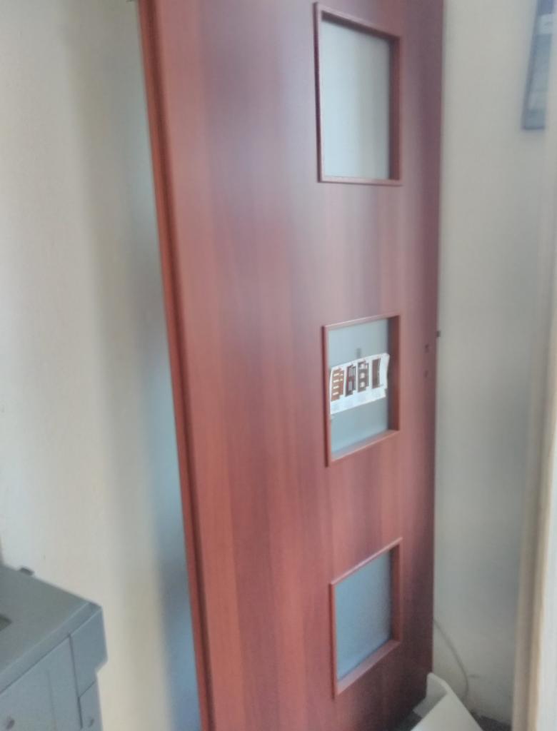 Brązowe drzwi wewnętrzne z trzema przeszkleniami, wykonane przez firmę z Zabrza, opierają się o ścianę w trakcie montażu.