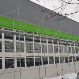 Ocieplanie elewacji budynku biurowego z szarymi ścianami i zielonym pasem, widoczne rusztowania i śnieg na ziemi.