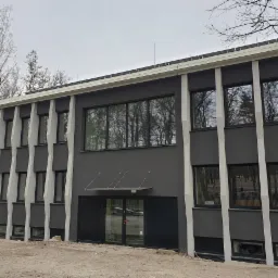 Dwukondygnacyjny budynek z szarą elewacją i białymi, ukośnymi elementami dekoracyjnymi, widoczne okna z odbiciami drzew, teren przed budynkiem w trakcie prac ziemnych.