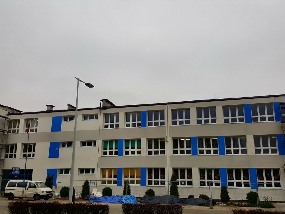 Szary budynek szkoły po remoncie elewacji z niebieskimi akcentami, widoczne rusztowania i zaparkowany biały bus.