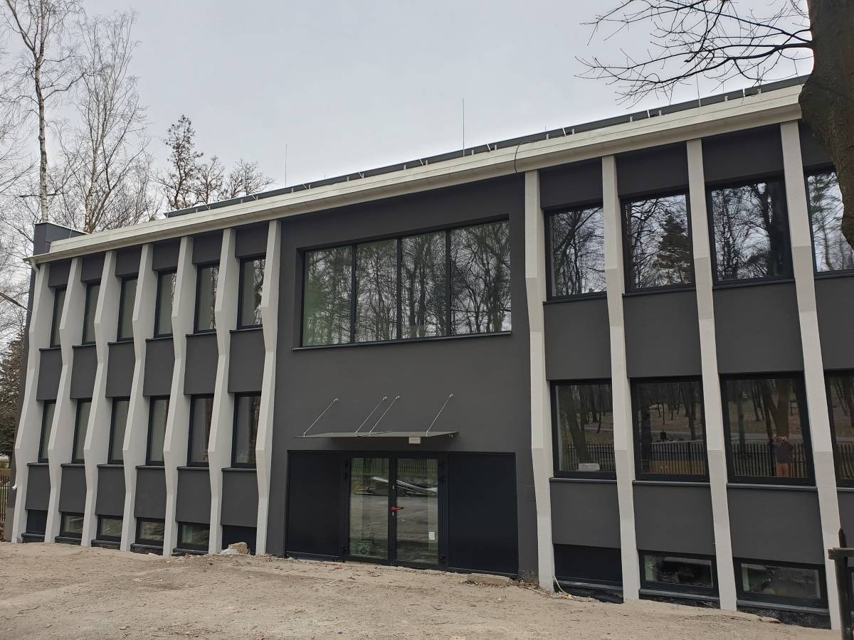 Dwukondygnacyjny budynek z szarą elewacją i białymi, ukośnymi elementami dekoracyjnymi, widoczne okna z odbiciami drzew, teren przed budynkiem w trakcie prac ziemnych.