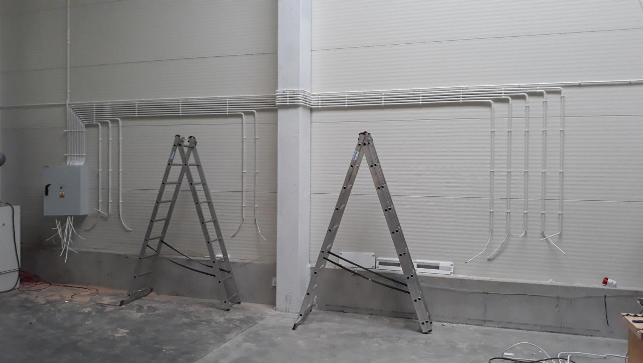 Szara skrzynka elektryczna z wystającymi kablami, drabiny aluminiowe i białe rury PCV ułożone równolegle na ścianie w hali.
