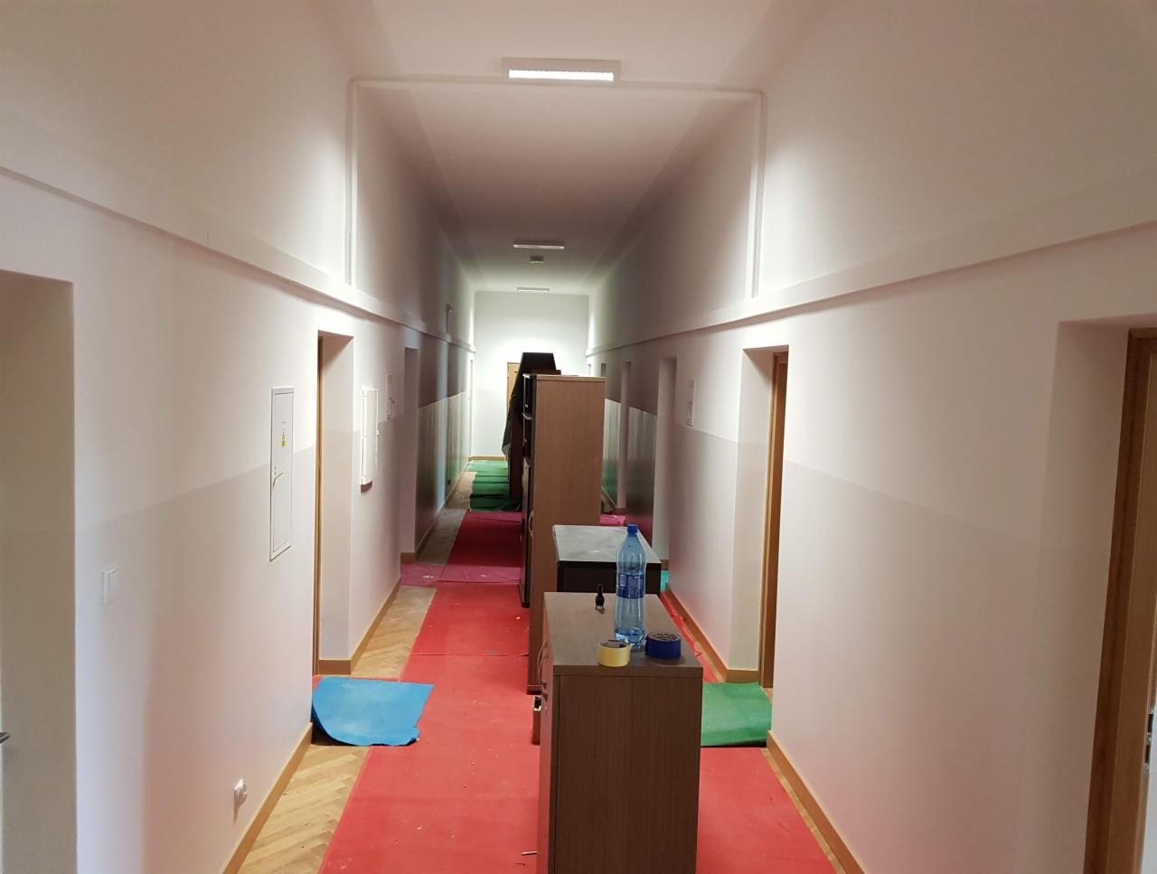 Korytarz biurowy w trakcie odświeżania: świeżo pomalowane ściany w odcieniach bieli, zabezpieczona podłoga czerwoną folią malarską, widoczne meble biurowe i butelka wody na szafce.