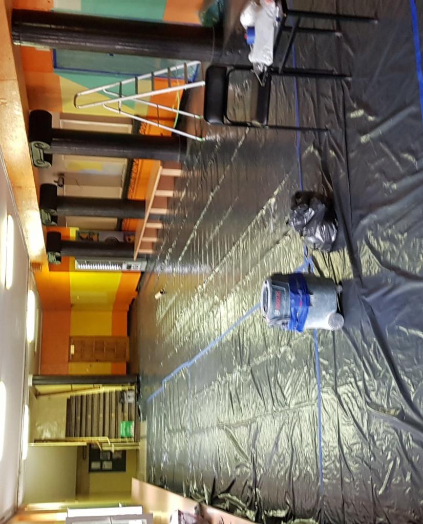 Przygotowanie do malowania dużej sali: podłoga zabezpieczona folią malarską, widoczny laser krzyżowy Bosch, drabina i krzesło w tle.