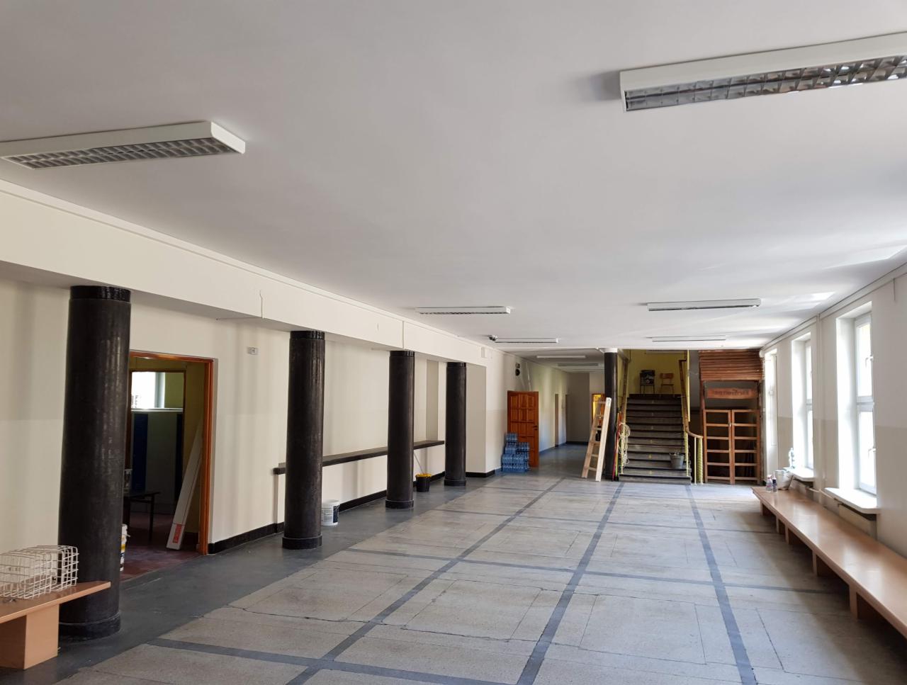 Długi korytarz biurowy z pomalowanymi na biało ścianami i sufitem, czarnymi kolumnami i szarą podłogą w kratkę, widoczne drzwi i schody w tle.