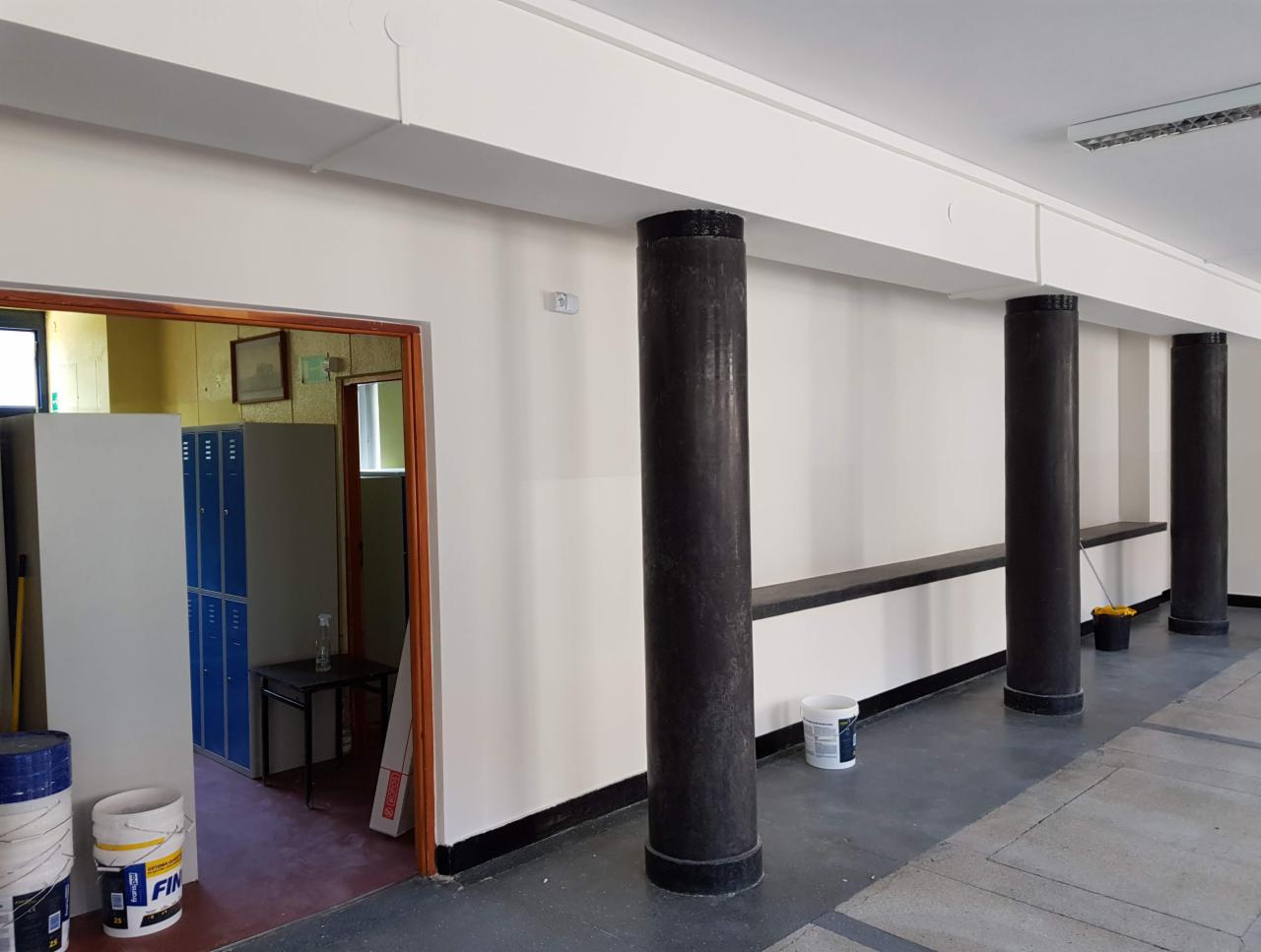 Korytarz biurowy po odświeżeniu ścian, widoczne pomalowane filary i wiaderka po farbie, w tle szatnia z niebieskimi szafkami.