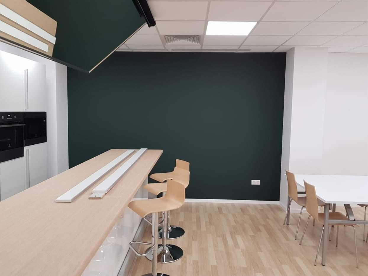 Nowoczesne biuro z ciemnozieloną ścianą po odświeżeniu, jasne drewniane podłogi, minimalistyczne meble i oświetlenie.