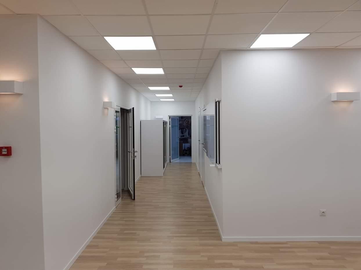 Długi, jasny korytarz biurowy po odświeżeniu, z białymi ścianami, drewnianą podłogą i nowoczesnym oświetleniem sufitowym oraz ściennymi lampami.