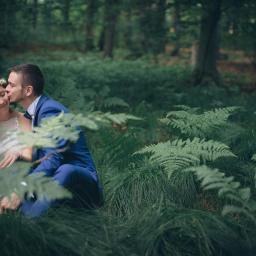 House of images - Panna młoda w wianku i pan młody w niebieskim garniturze siedzą w lesie, pan młody całuje pannę młodą w policzek, otoczeni paprociami.