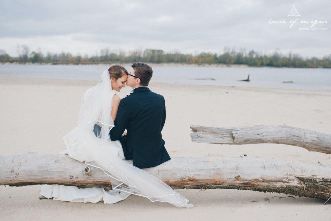 Para młoda siedząca na pniu drzewa na piaszczystej plaży, pan młody całuje pannę młodą w czoło, w tle woda i drzewa, pochmurne niebo.