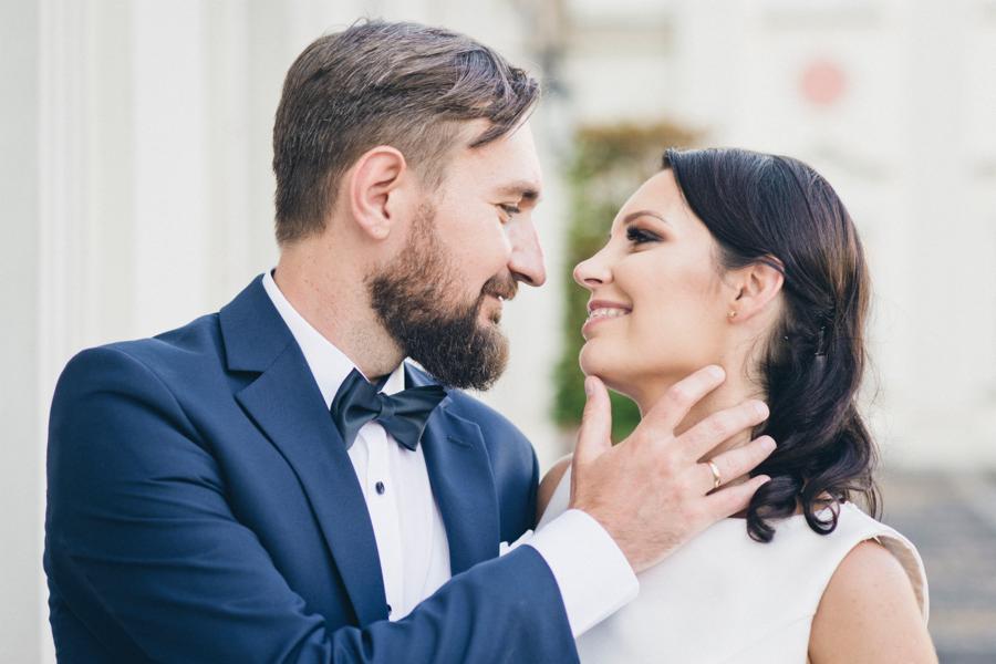 Uśmiechnięta para młoda patrzy na siebie, panna młoda dotykana przez pana młodego za podbródek, widoczne kolumny w tle.