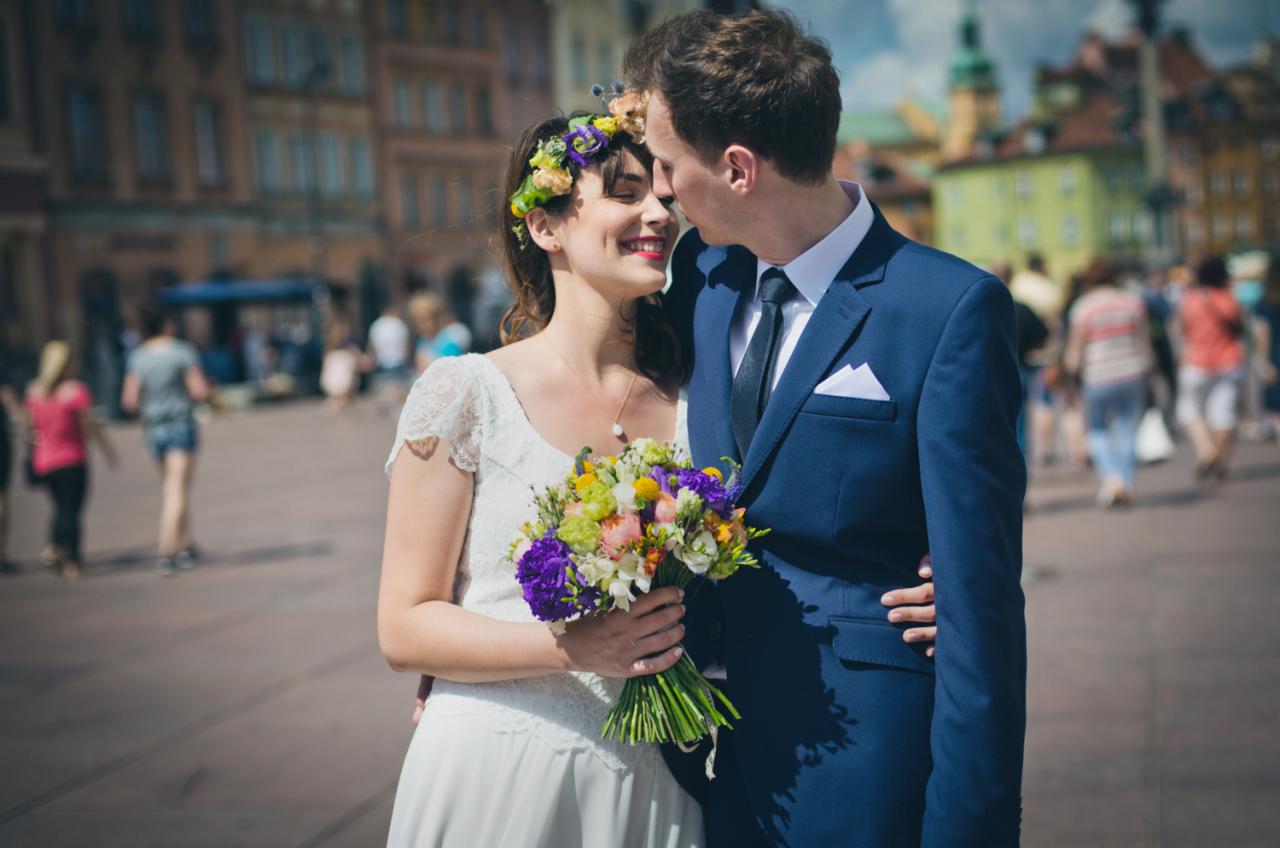 Uśmiechnięta panna młoda w wianku z kwiatów i koronkowej sukni, trzyma bukiet, wtulona w pana młodego w granatowym garniturze na tle Rynku Starego Miasta w Warszawie.