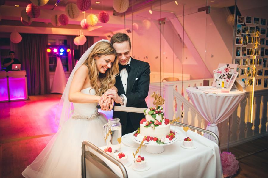 Para młoda uśmiechnięta kroi tort weselny udekorowany truskawkami i malinami, w tle dekoracje sali i DJ.