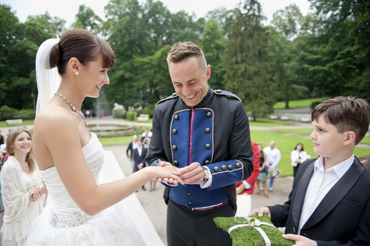 Panna młoda w białej sukni otrzymuje obrączkę od mężczyzny w galowym mundurze, chłopiec trzyma poduszkę na obrączki z mchu, w tle park i goście.