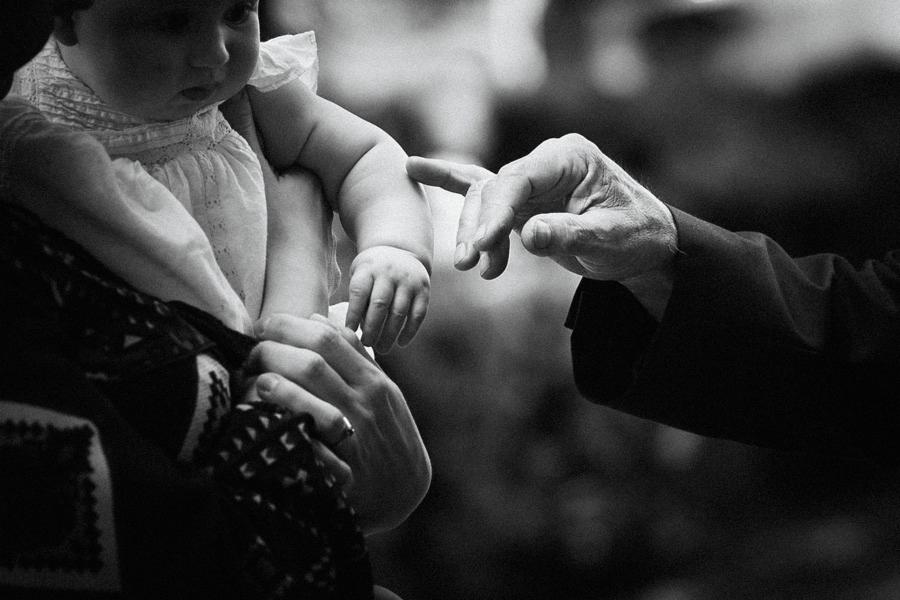 Czarno-białe zdjęcie niemowlęcia w białej sukience trzymanego na rękach, z dłonią osoby dorosłej dotykającej jego ramienia