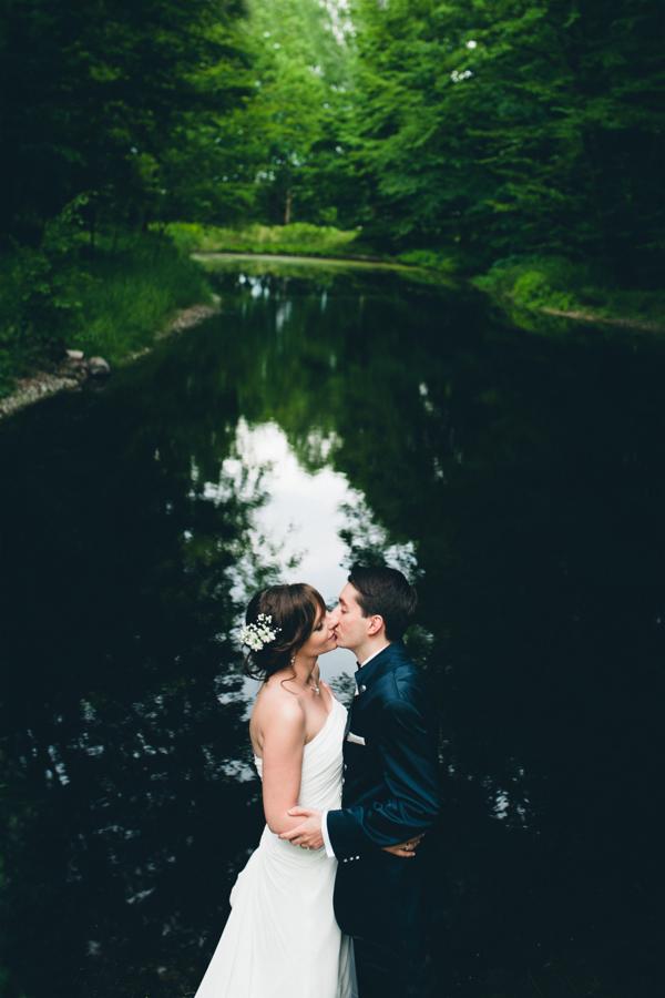 Pocałunek pary młodej na tle ciemnej wody i zieleni drzew, bukiet białych kwiatów we włosach panny młodej.