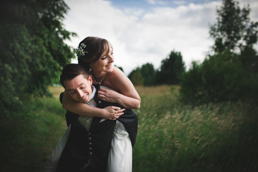 Uśmiechnięta panna młoda na barana u pana młodego na tle łąki i drzew w pochmurny dzień.