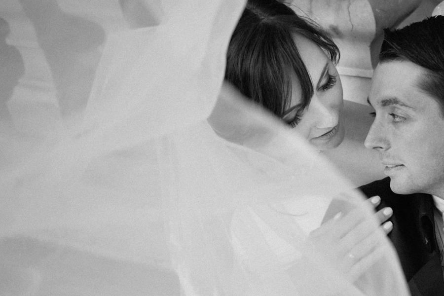 Czarno-białe zbliżenie pary młodej, panna młoda z ciemnymi włosami spogląda w dół na pana młodego, delikatna woalka w pierwszym planie, subtelne oświetlenie.