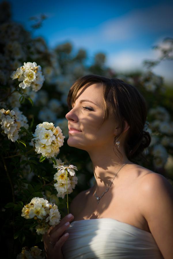 Panna młoda w białej sukni z odsłoniętymi ramionami, z zamkniętymi oczami, wącha białe róże w słoneczny dzień, delikatny cień na twarzy.