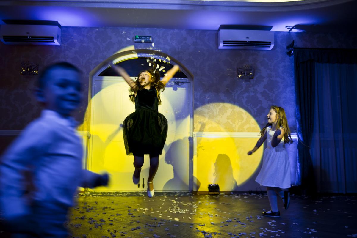 Dziewczynka w czarnej sukience skacze w górę, otoczona konfetti, podczas gdy inne dziecko w białej sukience biegnie obok, a chłopiec w koszuli jest rozmazany na pierwszym planie w sali...