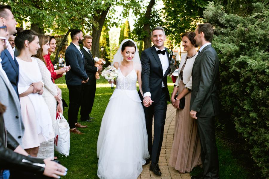 Para młoda, trzymając się za ręce, idzie ścieżką w otoczeniu gości weselnych w słoneczny dzień, panna młoda w białej sukni z bukietem, pan młody w czarnym smokingu z muchą.