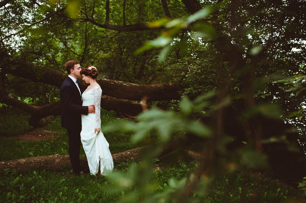 Para młoda w plenerze: pan młody w ciemnym garniturze całuje pannę młodą w koronkowej sukni na tle powalonych drzew w zalesionym otoczeniu.